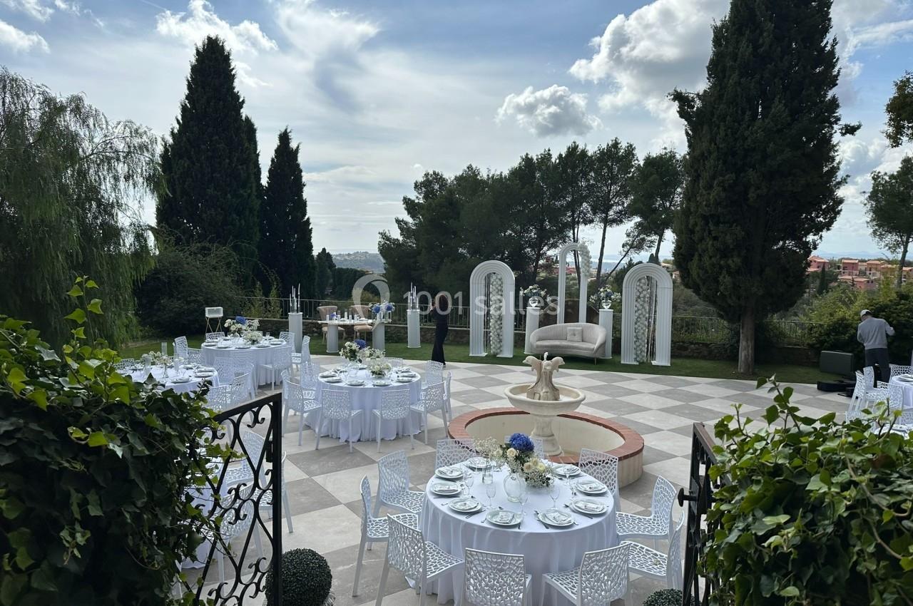 Terrasse extérieure aménagée pour un événement avec tables dressées, fontaine centrale et arches décoratives.