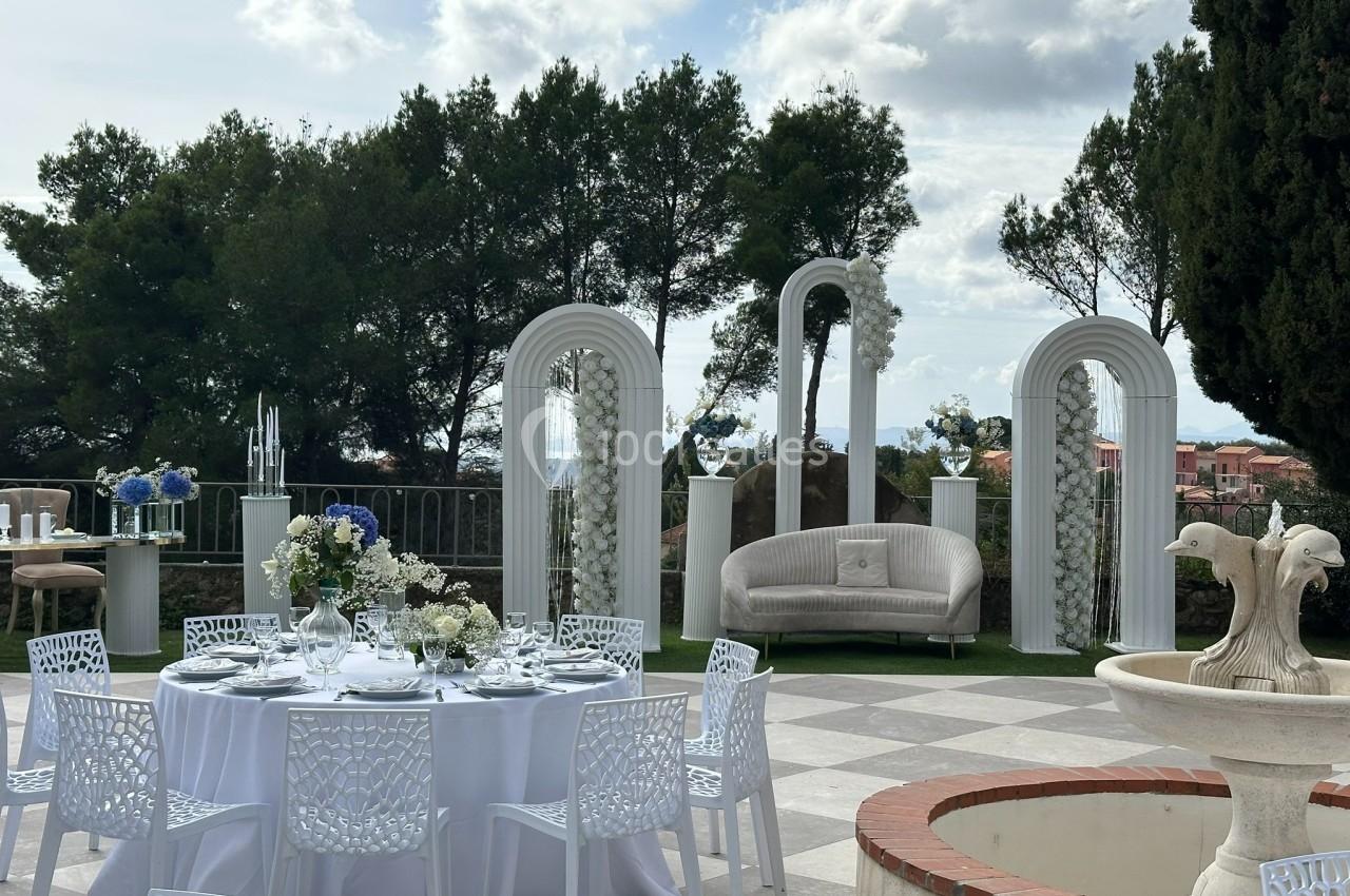 Décoration de mariage en extérieur avec tables dressées, arche florale blanche et vue sur des arbres et des bâtiments.