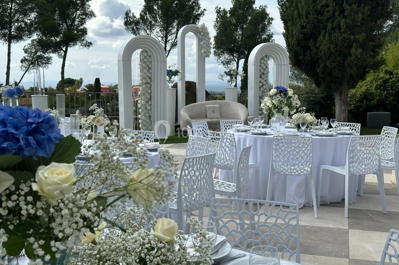 Tables élégamment dressées en extérieur avec des fleurs blanches et bleues, entourées d'arches décoratives et de verdure.