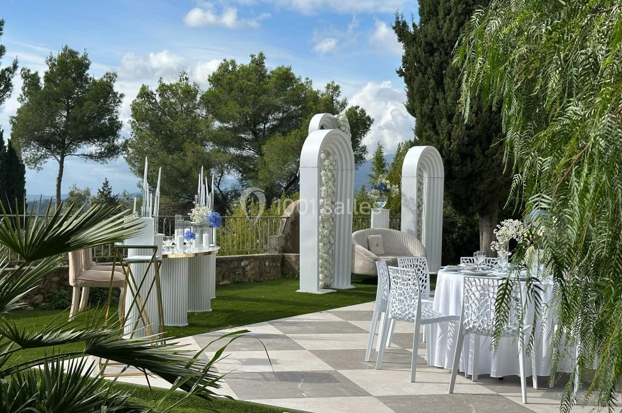 Décoration extérieure élégante pour un événement, avec arches blanches, table dressée et vue sur un jardin arboré.