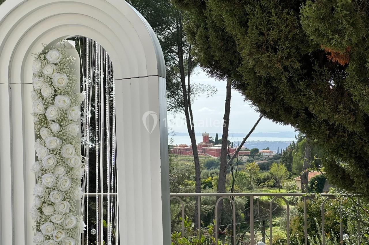Arche blanche décorée de roses blanches, avec vue sur un paysage verdoyant et une étendue d'eau en arrière-plan.