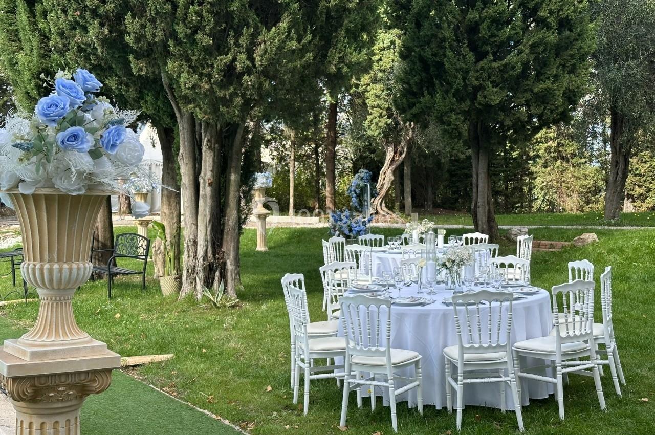 Table ronde décorée pour un événement, entourée de chaises blanches et située dans un jardin avec des arbres.