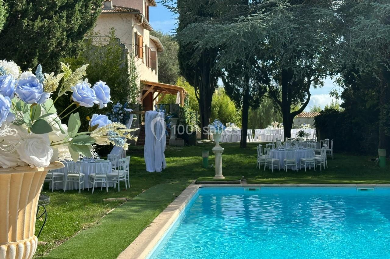 Jardin avec piscine, tables dressées pour un événement, décorations florales blanches et bleues, maison en arrière-plan.