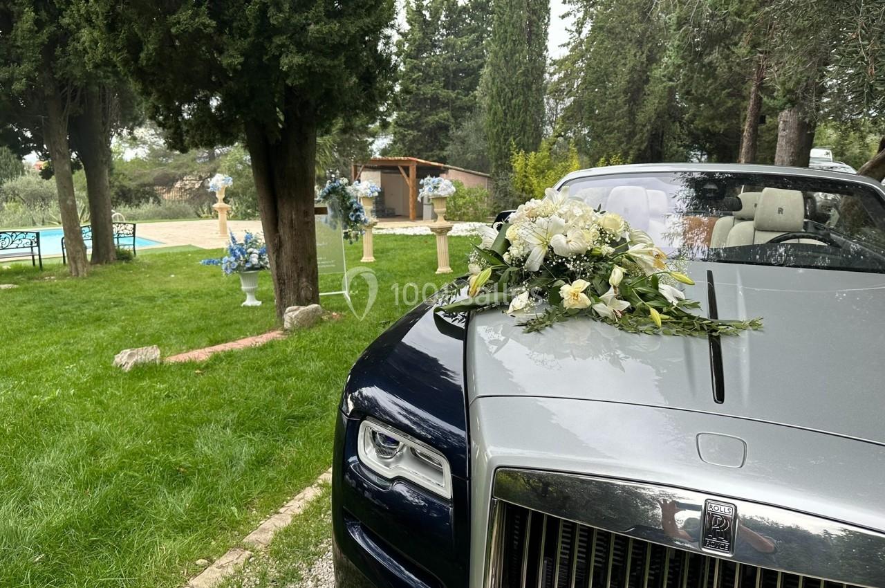 Voiture de luxe argentée avec un bouquet de fleurs blanches sur le capot, stationnée dans un jardin verdoyant.