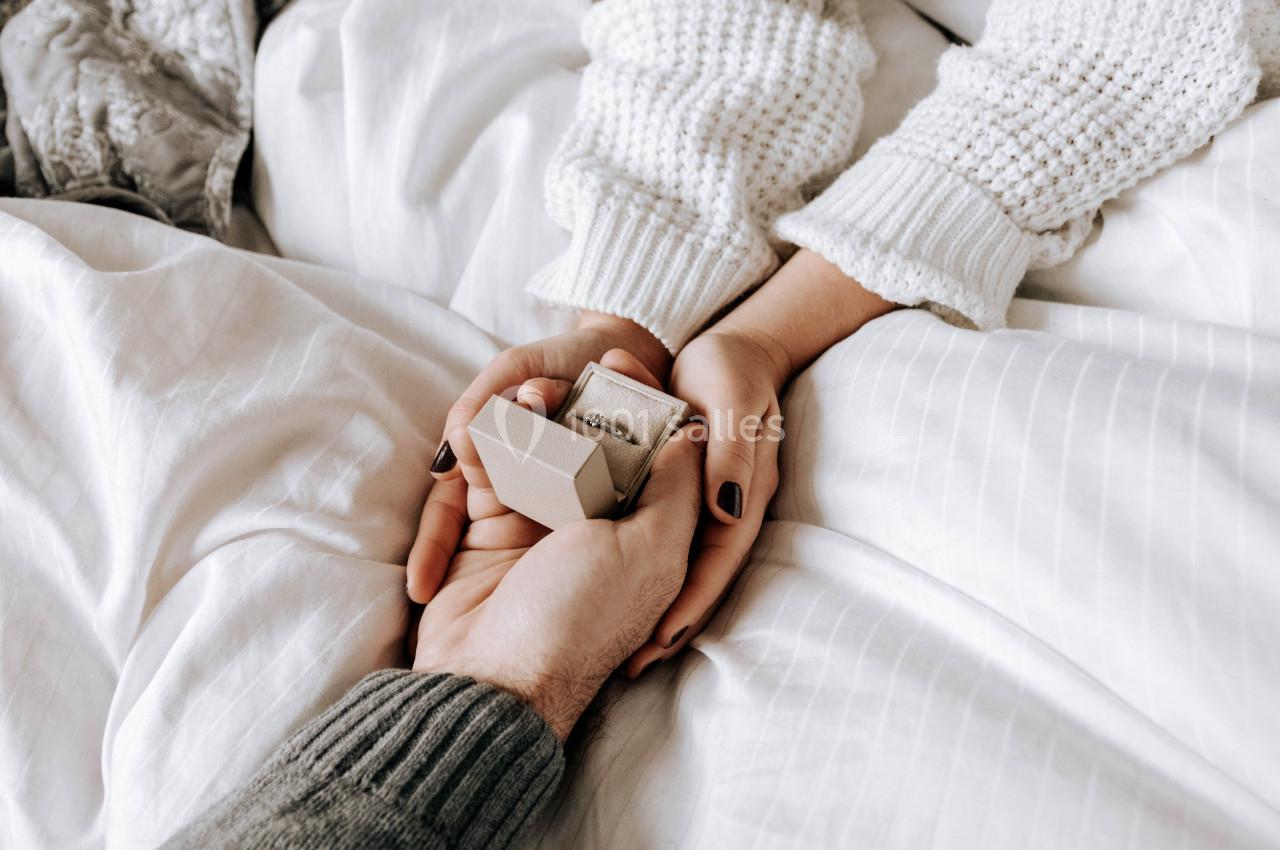 Une main tend une boîte contenant une bague à une autre main posée sur un lit aux draps blancs.
