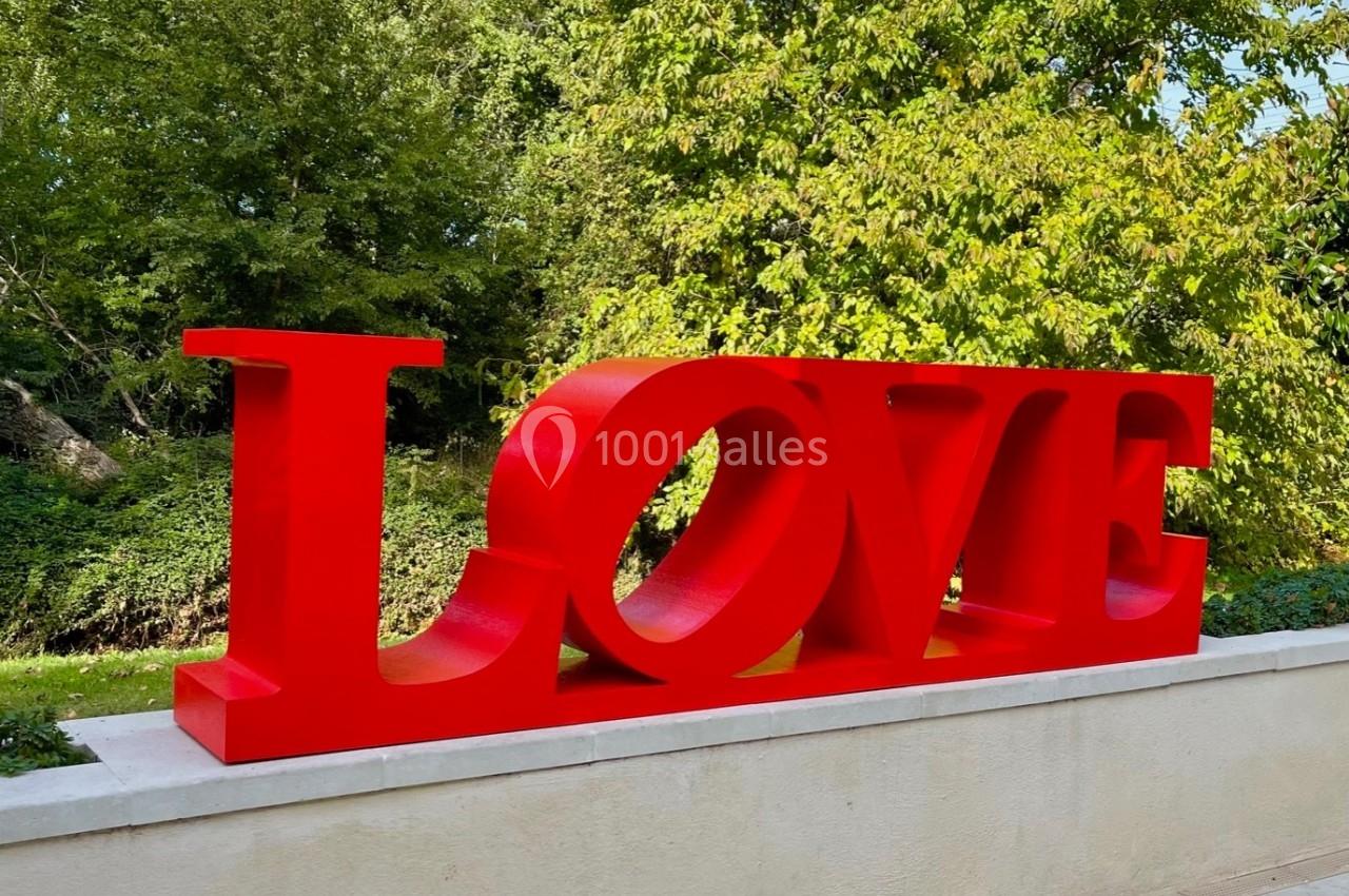 Sculpture rouge formant le mot ’LOVE’ posée sur un socle, avec des arbres en arrière-plan.