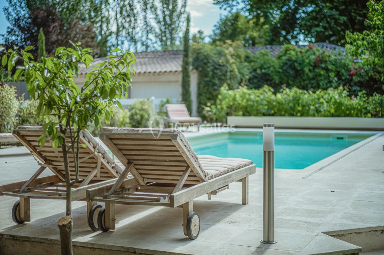 Deux transats en bois avec coussins près d'une piscine entourée de végétation et d'une maison en arrière-plan.