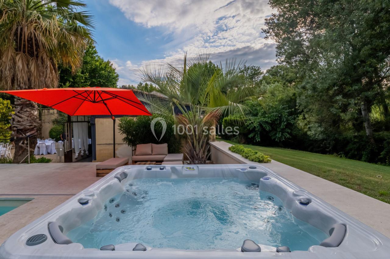 Vue d'un jacuzzi extérieur entouré de végétation, avec un parasol rouge et des meubles de jardin à proximité.