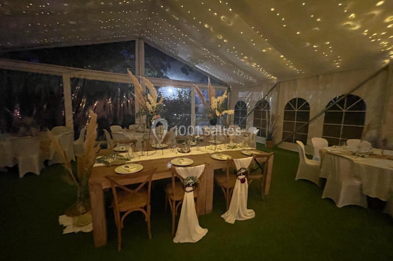 Salle de réception sous tente éclairée, avec tables décorées de fleurs et chaises habillées de tissu blanc.