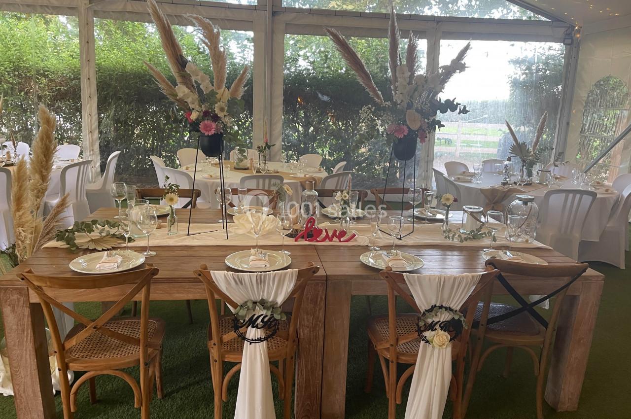 Table de mariage décorée avec des fleurs, des nappes blanches et des chaises ornées de voilages dans une salle lumineuse.
