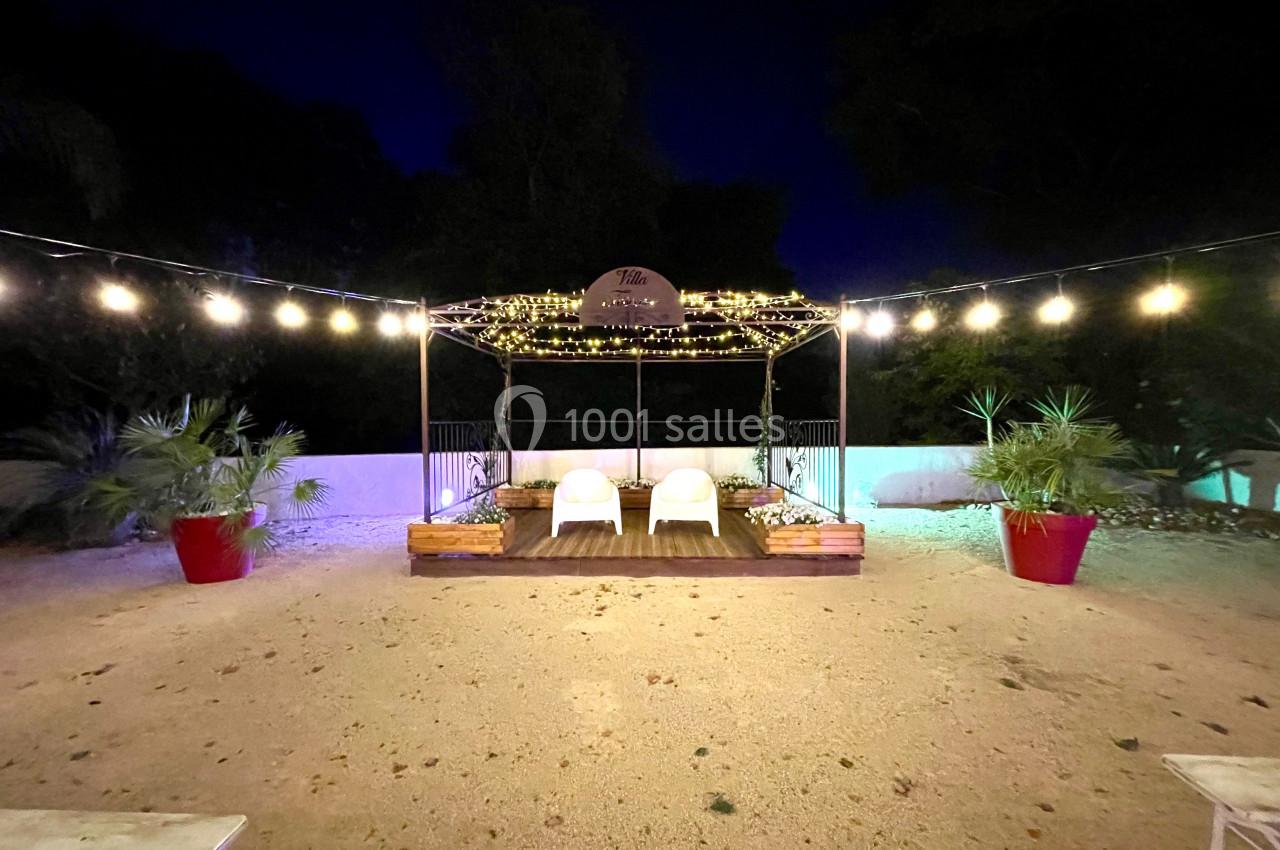 Scène extérieure éclairée la nuit avec guirlandes lumineuses, deux fauteuils blancs et plantes en pots rouges.