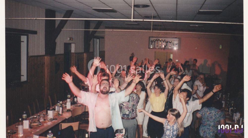 Des personnes dans une salle festive, levant les bras en l'air, entourées de tables avec des bouteilles et des verres.