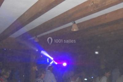 Salle sombre avec des poutres en bois, éclairée par des lumières colorées, des personnes discutent et se déplacent.