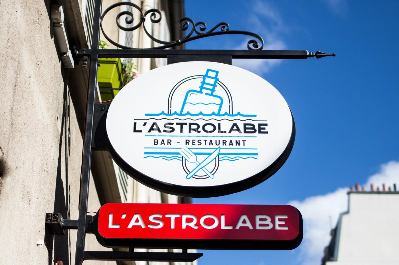 Enseigne d'un bar-restaurant nommé ’L'Astrolabe’ fixée sur une façade, avec un ciel bleu en arrière-plan.