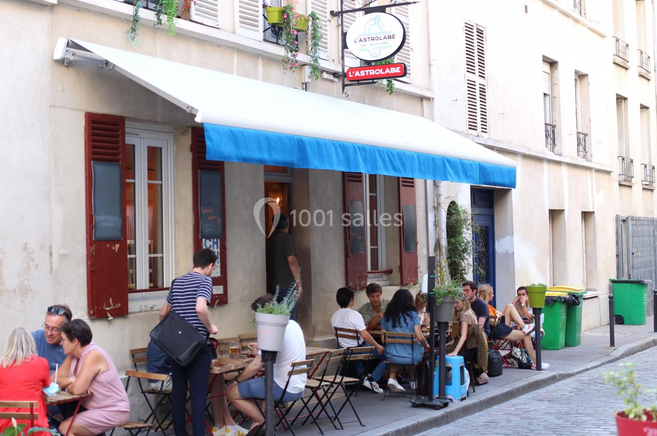 Terrasse d’un café animé avec des clients assis à des tables, située dans une rue calme bordée d’immeubles.