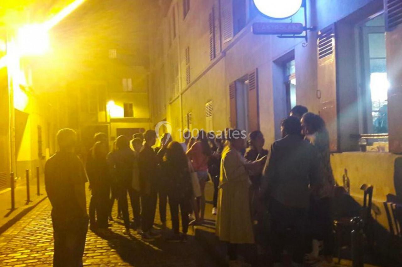 Groupe de personnes faisant la queue devant un bâtiment éclairé, dans une rue pavée en soirée.