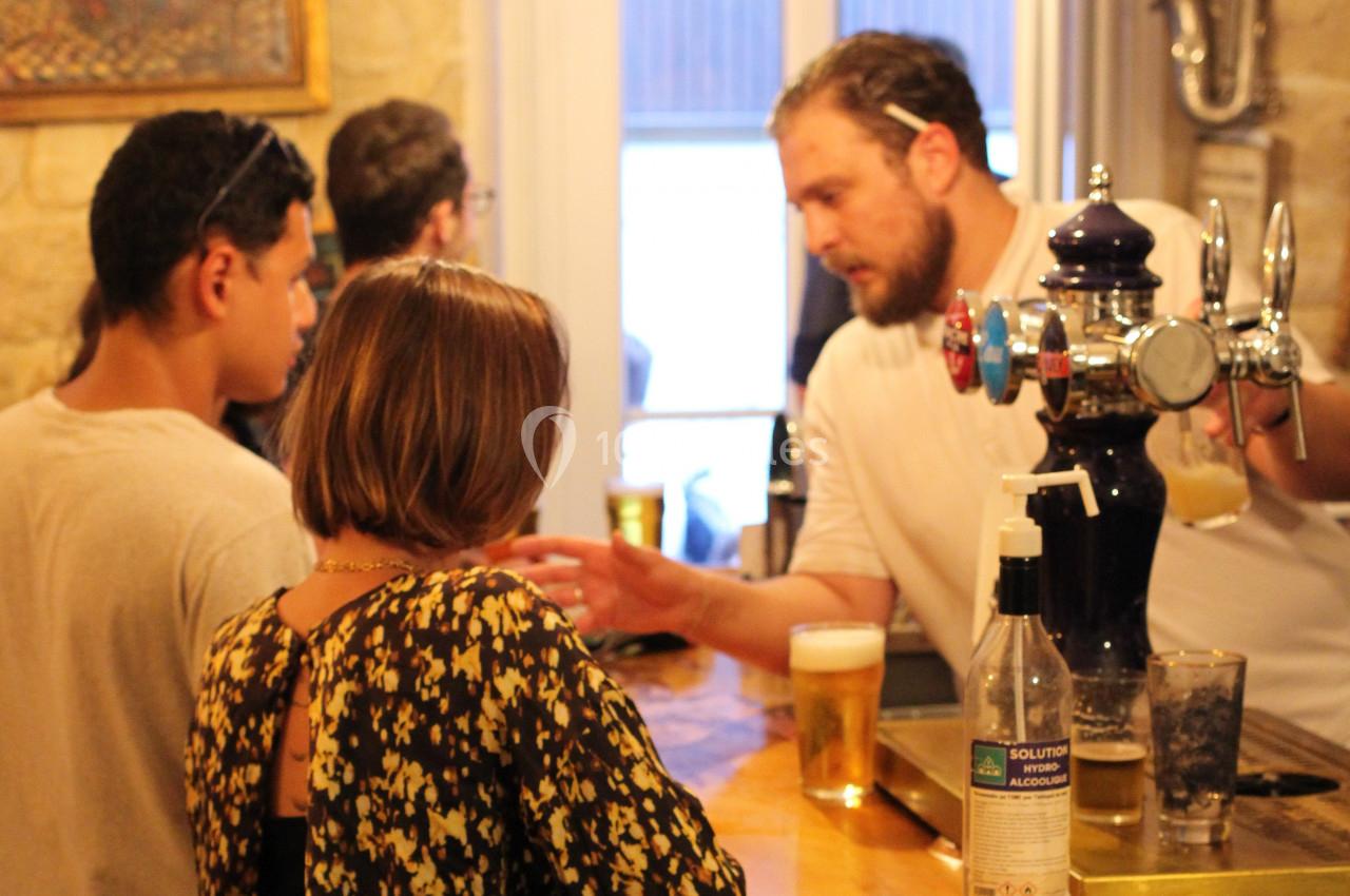 Un barman discute avec des clients autour d'un comptoir en bois, des verres de bière posés devant eux.