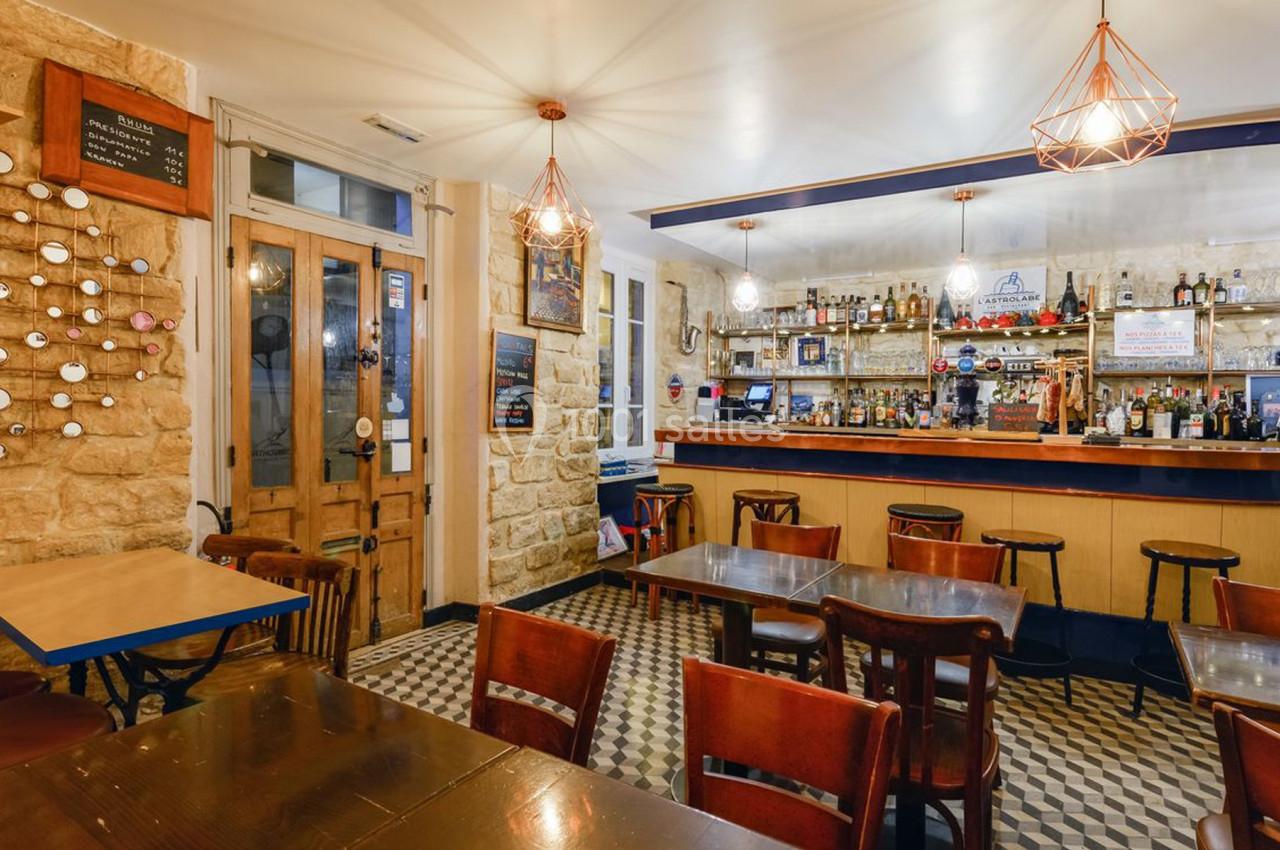 Intérieur d'un café avec tables en bois, chaises, bar en arrière-plan et décoration murale en pierre apparente.