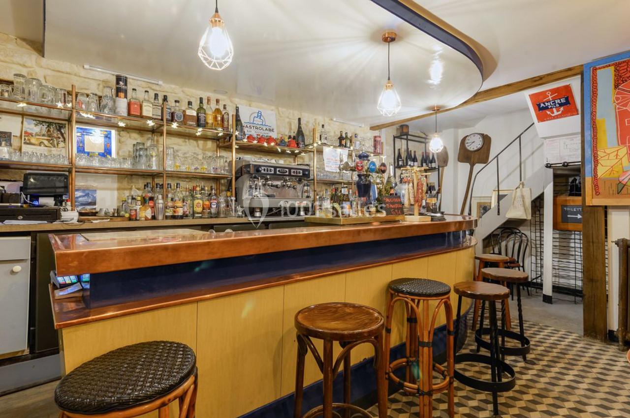 Bar avec comptoir en bois et cuivre, tabourets hauts, étagères remplies de bouteilles et éclairage suspendu.