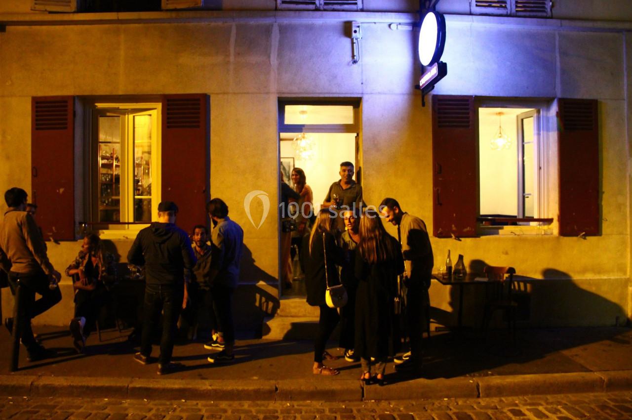 Groupe de personnes discutant devant un café éclairé en soirée, avec des fenêtres ouvertes et une lumière bleutée.