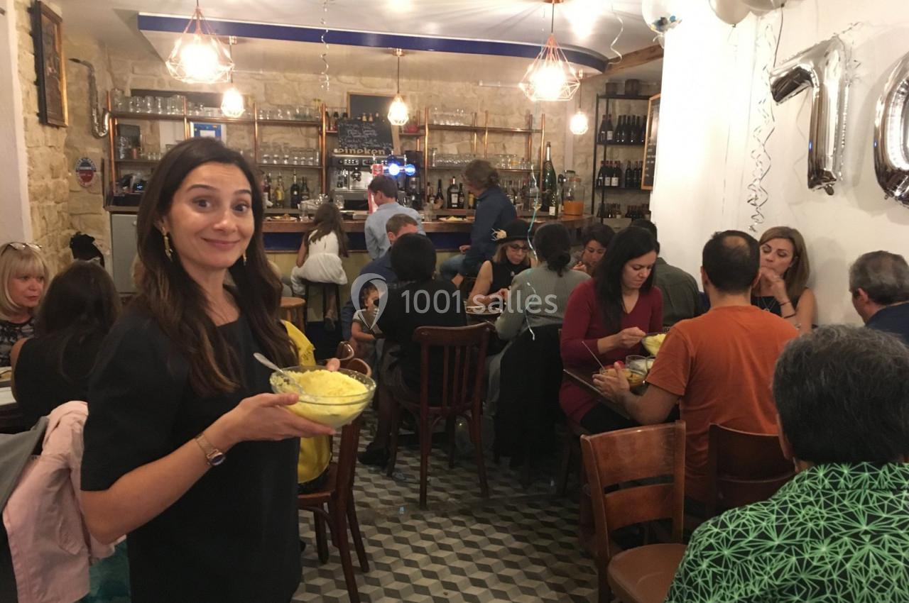 Une femme souriante tient un plat dans un restaurant animé avec des convives assis à des tables.