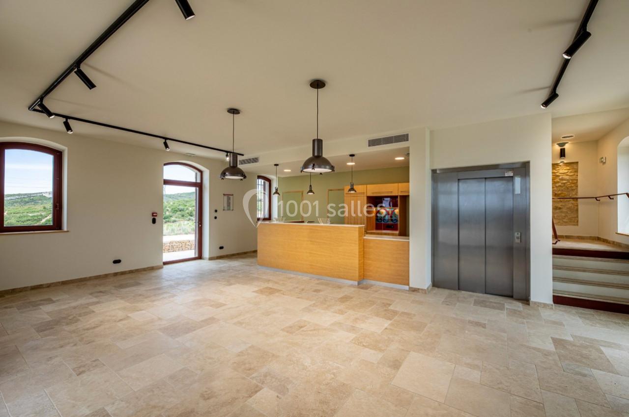 Salle lumineuse avec sol en pierre, comptoir en bois, ascenseur, éclairage suspendu et vue sur l'extérieur par des fenêtres.