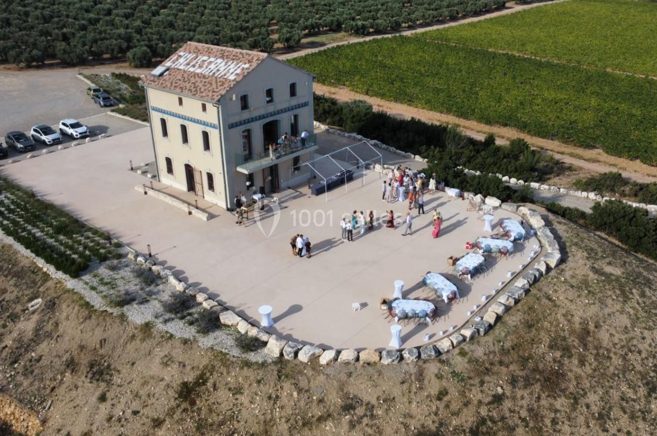 Vue aérienne d'une maison isolée entourée de champs, avec des invités rassemblés sur une terrasse pour un événement.