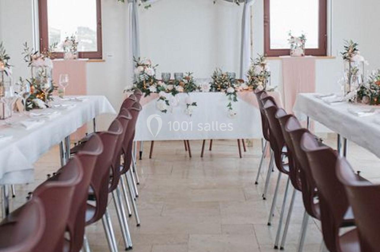 Salle décorée pour un mariage avec une arche florale, tables alignées et chaises bordeaux.