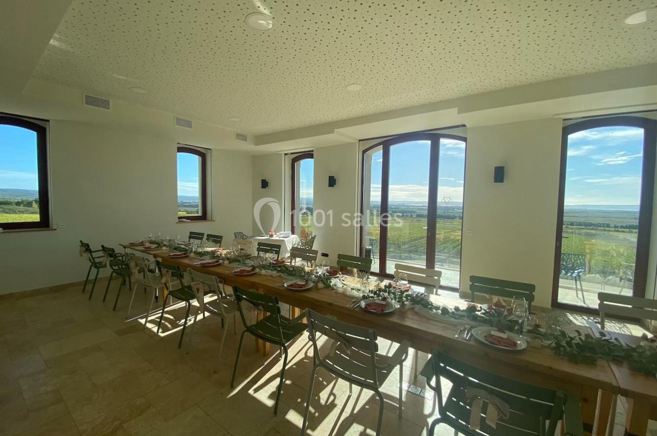 Salle lumineuse avec une grande table en bois décorée pour un repas, offrant une vue sur un paysage extérieur.