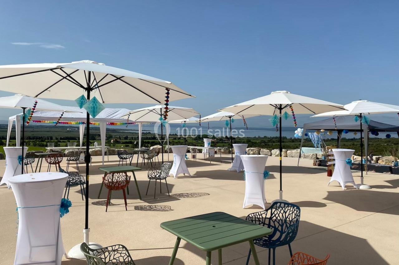 Terrasse ensoleillée avec tables, chaises colorées et parasols blancs, offrant une vue dégagée sur un paysage naturel.