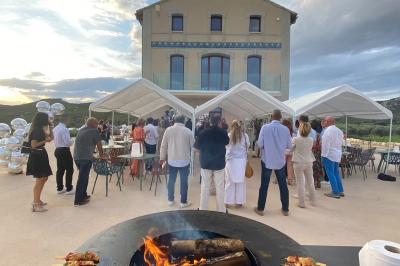 Personnes rassemblées devant une grande maison avec des tentes blanches, un brasero avec brochettes au premier plan.