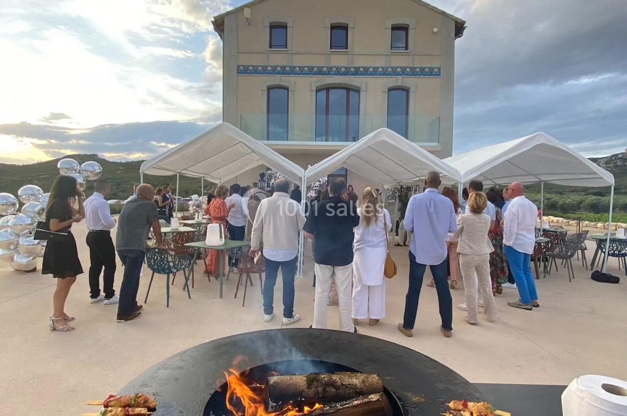 Personnes rassemblées devant une grande maison avec des tentes blanches, un brasero avec brochettes au premier plan.
