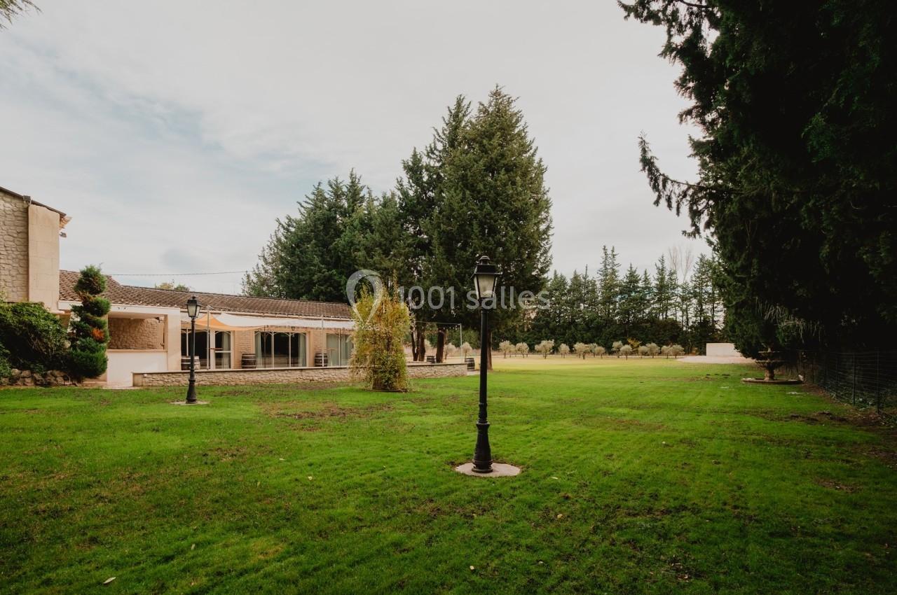 Jardin avec pelouse, lampadaires, arbres et une maison en arrière-plan sous un ciel nuageux.