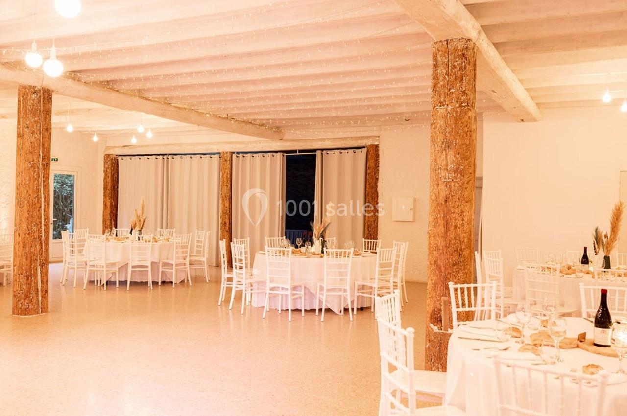 Salle de réception lumineuse avec tables rondes dressées, chaises blanches et colonnes en bois brut.