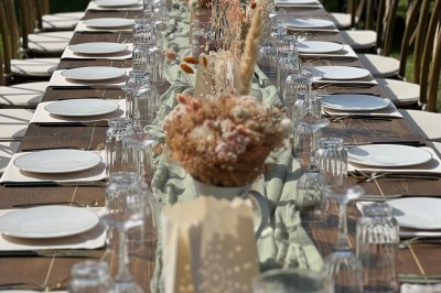 Table en bois dressée pour un repas, avec assiettes blanches, verres et décorations florales dans un cadre extérieur.
