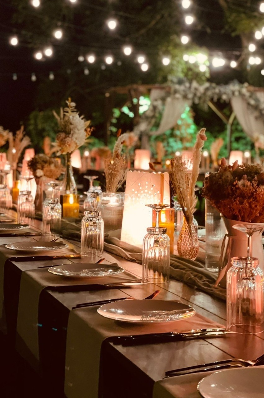 Table décorée pour un dîner en extérieur, éclairée par des guirlandes lumineuses et ornée de fleurs séchées.