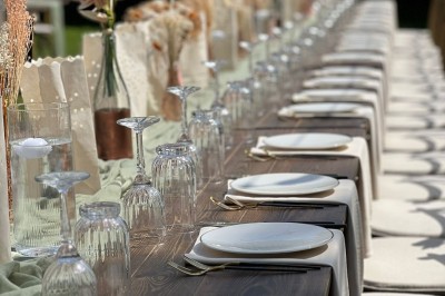 Table en bois dressée pour un repas, avec assiettes blanches, verres et décorations florales dans un cadre extérieur.