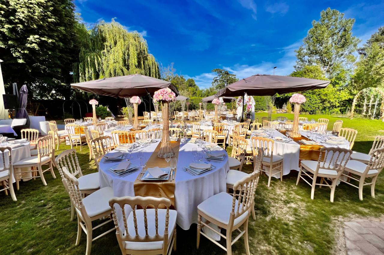 Tables rondes décorées pour un événement en plein air, entourées de chaises et disposées sous des parasols dans un jardin.