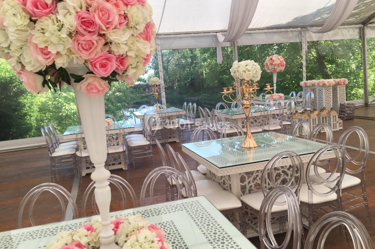 Salle décorée pour un événement avec des tables en verre, chaises transparentes et arrangements floraux roses et blancs.