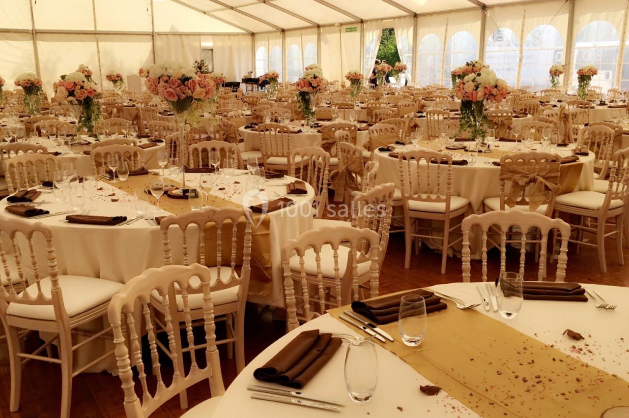 Salle de réception sous chapiteau avec tables rondes décorées de nappes blanches, chemins de table dorés et bouquets de…