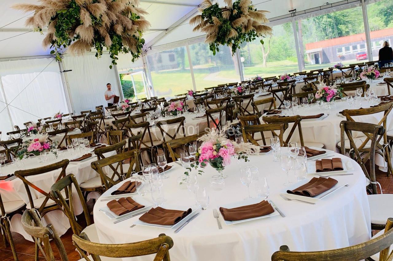 Salle de réception décorée avec des tables rondes dressées, nappes blanches, fleurs et chaises en bois.