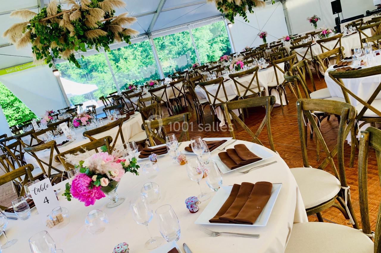 Salle de réception sous chapiteau avec tables dressées, nappes blanches, chaises en bois et décorations florales suspendues.