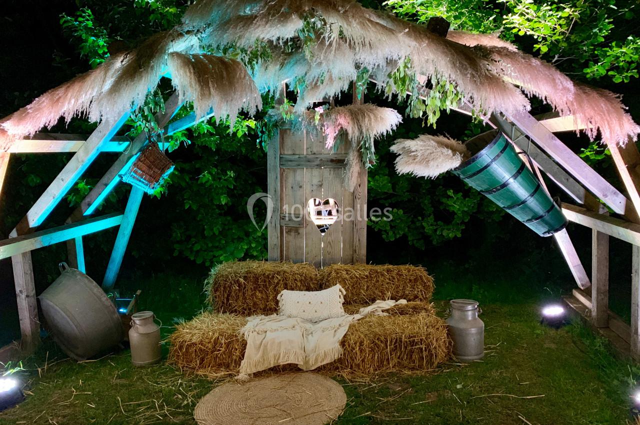 Structure en bois décorée de bottes de paille, de pampas et d'objets rustiques, éclairée par des lumières colorées.