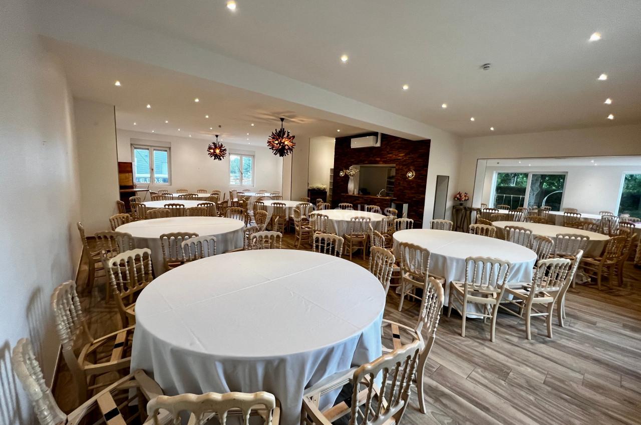 Salle lumineuse avec tables rondes dressées de nappes blanches et chaises en bois, éclairée par des suspensions modernes.