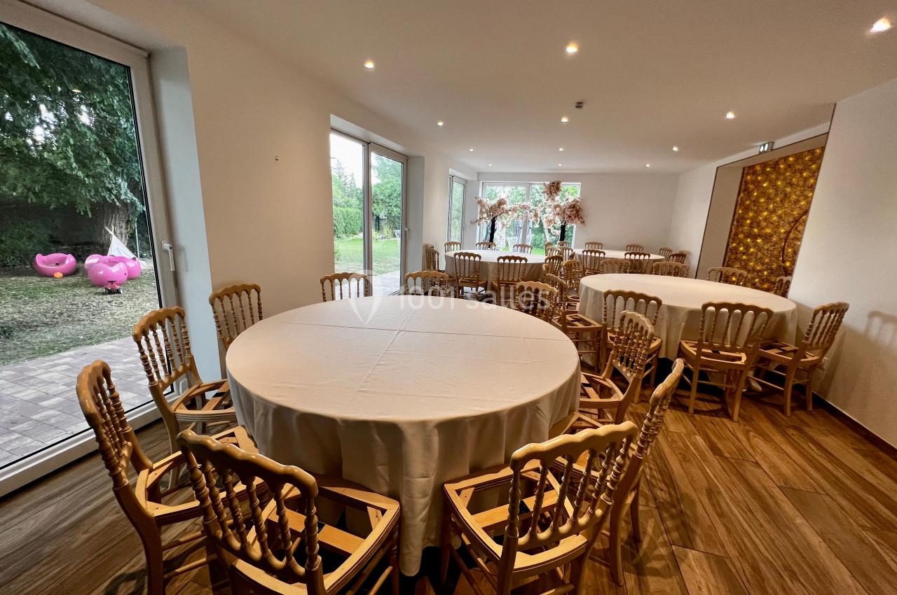 Salle lumineuse avec tables rondes dressées, chaises en bois, grandes baies vitrées et vue sur un jardin.