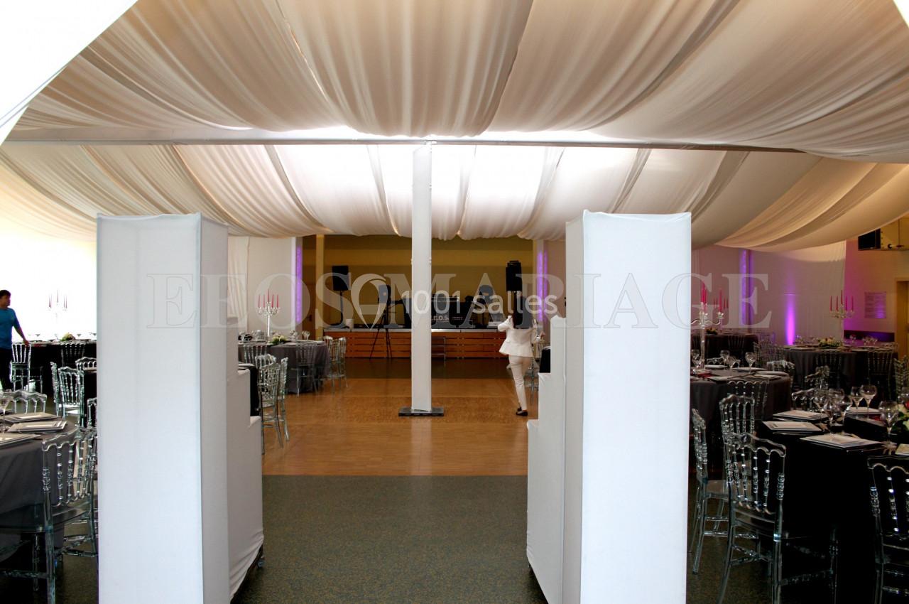 Salle de réception décorée avec des tables dressées, des chaises transparentes et un plafond drapé de tissu blanc.