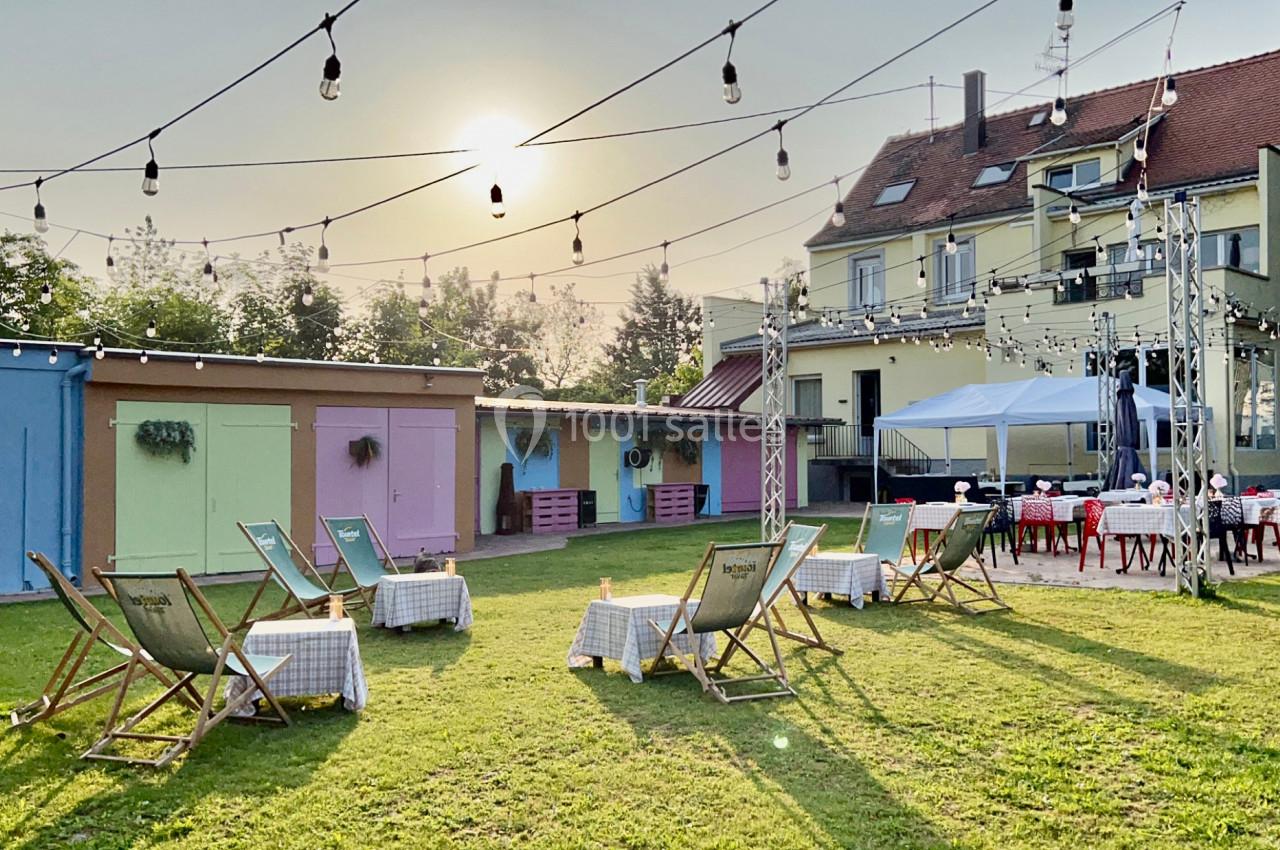 Chaises longues et tables disposées sur une pelouse, entourées de bâtiments colorés et éclairées par des guirlandes…