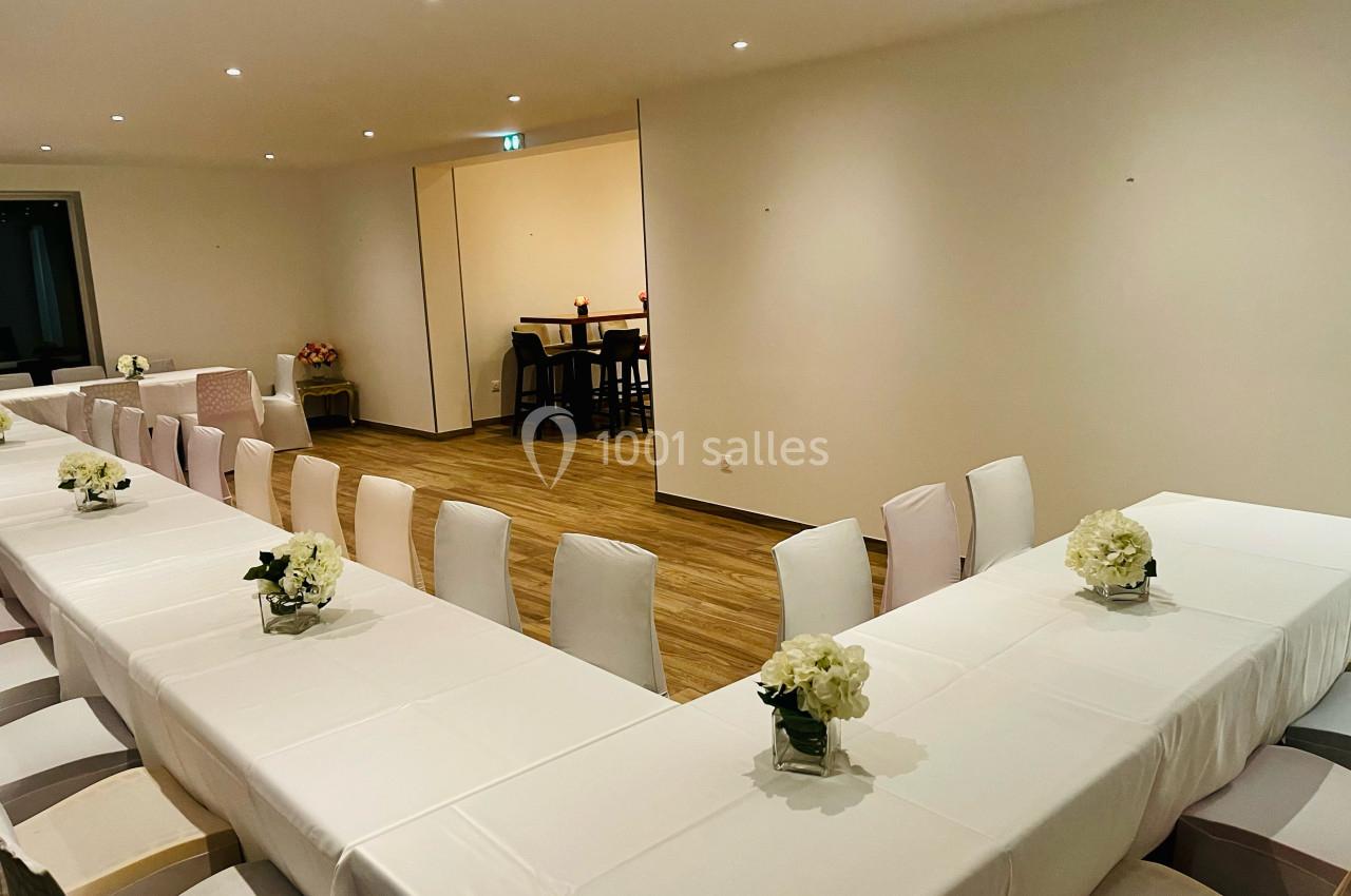 Salle de réception avec tables blanches en U, chaises recouvertes et décorations florales, éclairage doux.