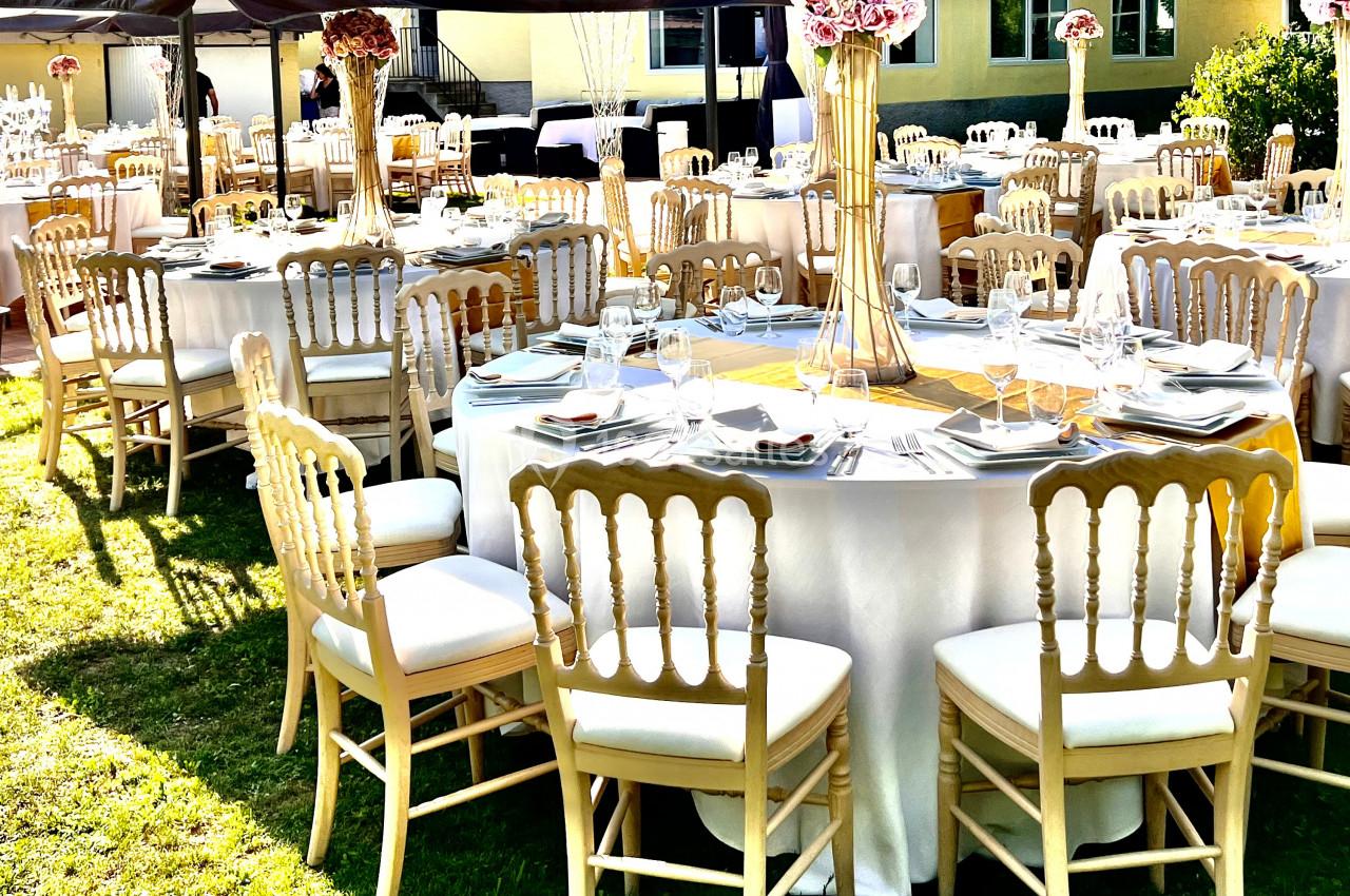 Tables rondes dressées avec nappes blanches, chaises en bois clair et décorations florales dans un jardin ensoleillé.