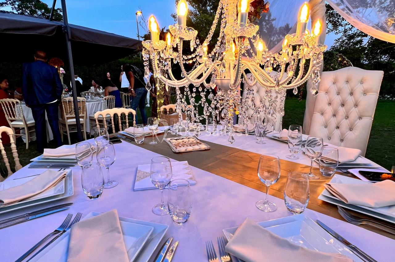 Table élégamment dressée en extérieur avec chandelier lumineux, vaisselle blanche et décor floral en arrière-plan.